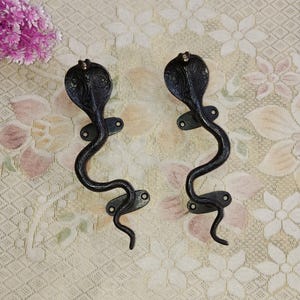 May include: A pair of black metal door handles shaped like cobras. The handles have a detailed design and are mounted on a white background.