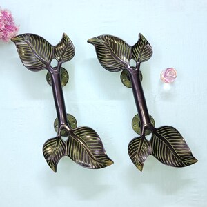 Peut inclure: Deux poignées de porte en forme de feuille avec une finition bronze. Les poignées présentent des veines de feuilles détaillées et une tige marron foncé. Elles sont montées sur des plaques rondes de couleur laiton. Une fleur rose et une rose décorative en verre sont en arrière-plan.