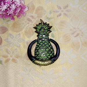 May include: A green and black pineapple door knocker with a gold ring. The knocker is made of metal and has a detailed design.