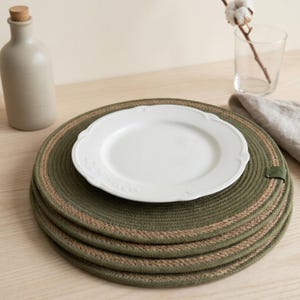 May include: A stack of four round, olive green braided rope placemats with a natural jute-colored border and a small green fabric tag on the edge. A single white ceramic plate with a decorative embossed rim rests on top of the placemats.