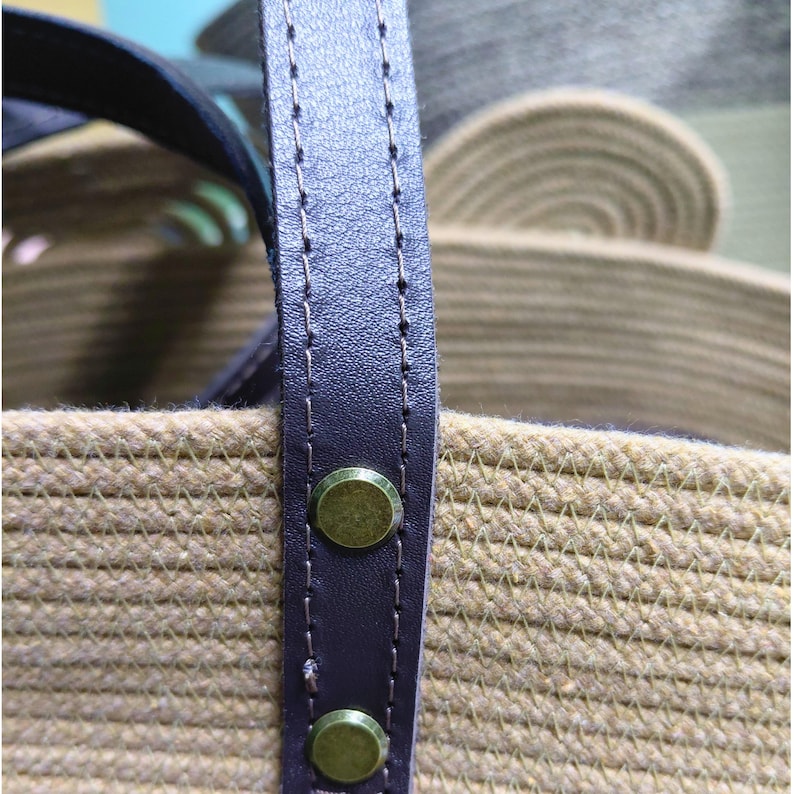 May include: Close-up of a woven beige tote bag with a dark brown leather handle. The handle is secured with brass-colored rivets. The bag's texture and color suggest a natural, casual style, suitable for everyday use.