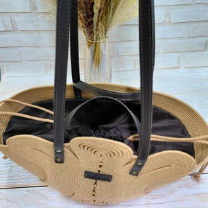 May include: A beige rope tote bag with black leather handles and accents. The bag has a unique, open design with a black interior and a small black label. The bag is sitting on a light wood surface.
