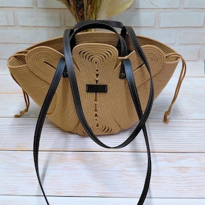 May include: A tan rope tote bag with black leather handles and a shoulder strap. The bag has a unique design with a central decorative element and a small black label. The bag is sitting on a light wood surface.