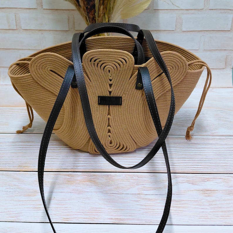 May include: A tan rope tote bag with black leather handles and a shoulder strap. The bag has a unique design with a central decorative element and a small black label. The bag is sitting on a light wood surface.