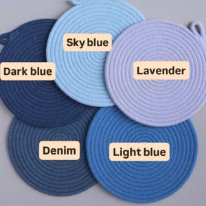 May include: A collection of five round, woven trivets in shades of blue and lavender. The trivets are labeled with the colors: Dark blue, Sky blue, Lavender, Denim, and Light blue. Each trivet has a loop for hanging.