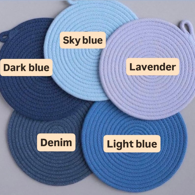 May include: A collection of five round, woven trivets in shades of blue and lavender. The trivets are labeled with the colors: Dark blue, Sky blue, Lavender, Denim, and Light blue. Each trivet has a loop for hanging.