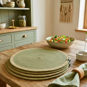 Peut inclure: Quatre sets de table ronds verts avec un motif en spirale et une bordure beige sont empilés sur une table en bois. Un saladier, une serviette avec un anneau en bois et un petit bol de sel sont également sur la table. Un meuble vert est en arrière-plan.