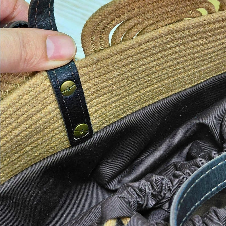 May include: Close-up of a woven straw handbag with a brown leather handle and accents. The bag features a dark interior and is held by a hand. The handle is secured with brass-colored rivets.