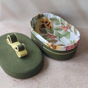 May include: An oval storage basket made of coiled olive green rope with a matching lid featuring a decorative yellow and green bow. The interior is lined with white fabric printed with a pattern of yellow flowers, green leaves, and small pink berries.