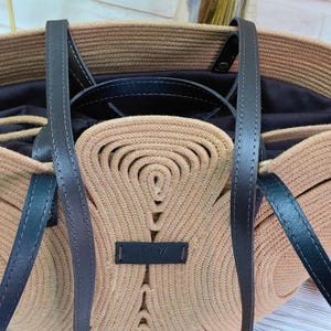 May include: A beige tote bag with black leather handles and trim. The bag is made of woven material and features a decorative design on the front. The interior of the bag is dark, and the handles are attached with metal hardware.