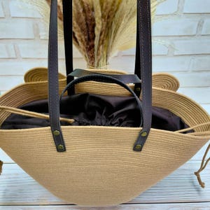May include: A beige woven tote bag with dark brown leather handles and accents. The bag has a drawstring closure and a dark brown interior. The bag is made of a woven material and has a unique shape.