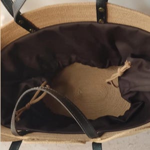 May include: A woven straw tote bag with black leather handles and a dark brown interior. The bag has a drawstring closure and is designed for carrying items. The bag is a neutral color and has a casual style.