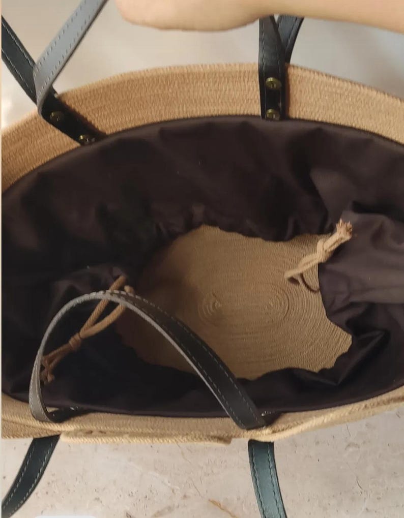 May include: A woven straw tote bag with black leather handles and a dark brown interior. The bag has a drawstring closure and is designed for carrying items. The bag is a neutral color and has a casual style.