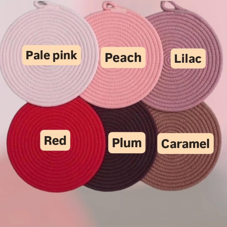 May include: Six round, woven trivets in various colors: pale pink, peach, lilac, red, plum, and caramel. Each trivet has a loop for hanging and a label with its color name.