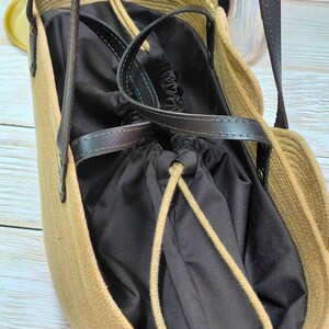 May include: A beige tote bag with a dark brown interior and leather handles. The bag has a drawstring closure and a woven texture. The handles are attached with metal rivets.