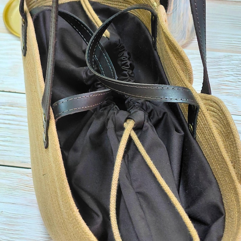 May include: A beige tote bag with a dark brown interior and leather handles. The bag has a drawstring closure and a woven texture. The handles are attached with metal rivets.
