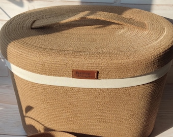 Puede incluir: Una cesta de almacenamiento redonda y con tapa hecha de cuerda trenzada de color marrón claro. Una banda blanca rodea la cesta, y una pequeña etiqueta marrón con texto está fijada en la parte delantera. La cesta está diseñada para la organización del hogar.