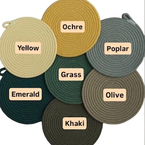 May include: A collection of seven round, woven trivets in various colors: yellow, ochre, emerald, grass, poplar, olive, and khaki. Each trivet has a loop for hanging and a label with its color name.