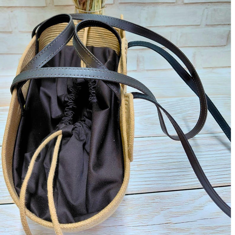 May include: A beige and brown tote bag with a black interior and black leather straps. The bag has a drawstring closure and a woven design. The bag is sitting on a light wood surface.