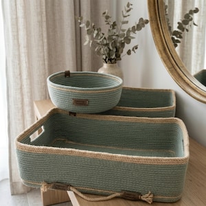 May include: A set of sage green rope storage baskets featuring a rectangular tray with integrated handles and a round bowl. Both items are constructed from coiled rope with tan accents and include small brown leather labels with text.