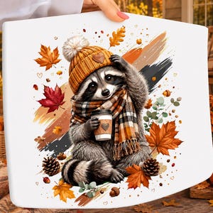 Könnte beinhalten: Eine weiße Oberfläche mit einer grafischen Illustration eines Waschbären, der eine senfgelbe Strickmütze mit Bommel und einen karierten Schal trägt. Der Waschbär hält einen Kaffeebecher. Das Motiv ist von Herbstblättern und Eicheln umgeben.