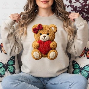 May include: A tan sweatshirt featuring a 3D teddy bear design. The bear is brown with a red heart and bow. The sweatshirt is worn by a person sitting on a couch with butterfly pillows.