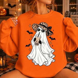 May include: An orange crewneck sweatshirt featuring a graphic of a white ghost wearing a leopard print sun hat and black sunglasses. The ghost holds a plastic cup of boba tea. The design includes small black bats and four-pointed star shapes.