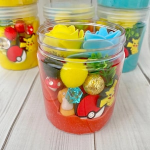 Op de afbeelding: Een doorzichtige plastic pot gevuld met rode slijm, voorzien van diverse kleine plastic bedeltjes waaronder een gele bloem, een blauwe bloem, een gele pompon, een gouden glitterbal, een blauw kristal, een rood-witte Pok&eacute; Ball en een figuurtje.
