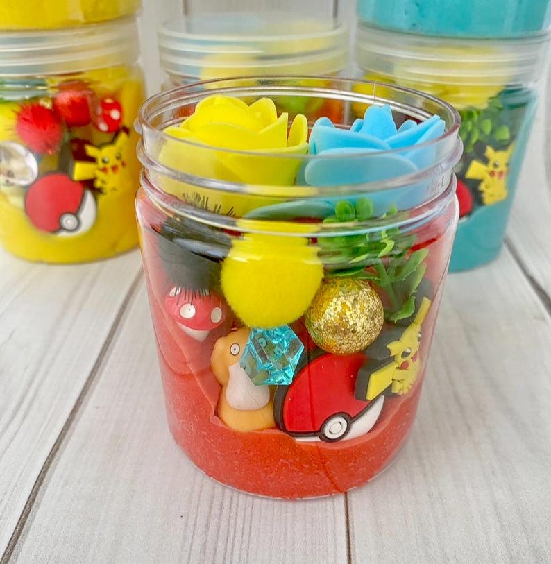Op de afbeelding: Een doorzichtige plastic pot gevuld met rode slijm, voorzien van diverse kleine plastic bedeltjes waaronder een gele bloem, een blauwe bloem, een gele pompon, een gouden glitterbal, een blauw kristal, een rood-witte Pok&eacute; Ball en een figuurtje.