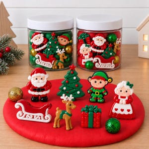 May include: A collection of Christmas-themed flatback resin cabochons including Santa Claus, an elf, a reindeer, a sleigh, a Christmas tree, a gift box, and Mrs. Claus, displayed on a red circular mat with two clear jars filled with similar pieces.