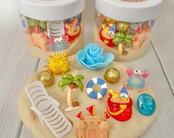 May include: Two clear plastic jars with white lids labeled Beach Play Dough Jar containing beige dough and beach-themed charms. A flat beige dough base features a white lounge chair, palm tree, sandcastle, red bucket, blue life ring, and crab.