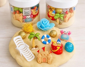 Könnte beinhalten: Zwei durchsichtige Kunststoffdosen mit weißen Deckeln und der Aufschrift Beach Play Dough Kit enthalten beigefarbenen Teig und Miniaturfiguren zum Thema Strand. Eine flache Scheibe aus beigem Teig ist mit einem weißen Liegestuhl, einer Palme, einer Sandburg, einem roten Eimer, einem blauen Rettungsring, einer Krabbe und einer blauen Blume verziert.