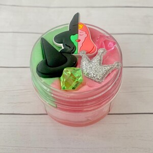 Peut inclure: Un récipient en plastique transparent rempli de slime rose et vert. Le slime est décoré d'un chapeau de sorcière noir, d'une gemme verte, d'une couronne à paillettes argentées et d'un visage divisé de sorcière et de princesse.