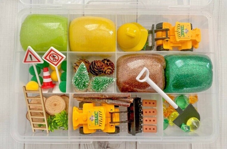 Puede incluir: Un recipiente de pl&aacute;stico transparente lleno de juguetes y plastilina con tem&aacute;tica de construcci&oacute;n. Incluye un casco amarillo, camiones de juguete, se&ntilde;ales de tr&aacute;fico, &aacute;rboles en miniatura y plastilina verde, amarilla, marr&oacute;n y verde brillante.
