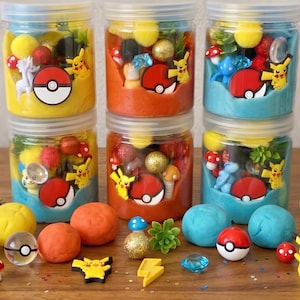 Op de afbeelding: Zes doorzichtige plastic potten gevuld met kleurrijke boetseerklei en diverse kleine plastic figuurtjes, waaronder Pikachu en Pok&eacute; Balls. De potten staan in twee rijen, met extra losse kleiballen, miniatuurpaddenstoelen en bedeltjes verspreid aan de voorzijde.
