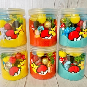 Op de afbeelding: Vijf doorzichtige plastic potjes gevuld met gekleurde boetseerklei, elk met kleine plastic figuurtjes waaronder gele Pikachu-personages, rood-witte Pok&eacute; Ball-fiches, glinsterende bollen en diverse decoratieve miniatuurvoorwerpen.