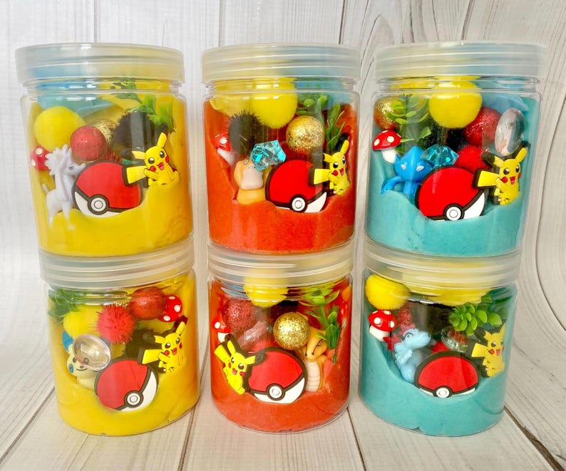 Op de afbeelding: Vijf doorzichtige plastic potjes gevuld met gekleurde boetseerklei, elk met kleine plastic figuurtjes waaronder gele Pikachu-personages, rood-witte Pok&eacute; Ball-fiches, glinsterende bollen en diverse decoratieve miniatuurvoorwerpen.