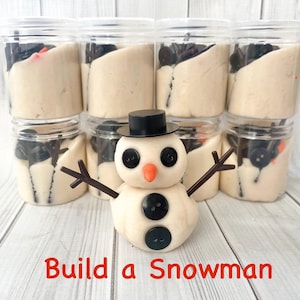 Peut inclure: Un kit de bricolage sur le thème du bonhomme de neige avec des bocaux en plastique transparent remplis de pâte blanche et d'accents noirs. Un petit bonhomme de neige avec un chapeau noir, des yeux en bouton et un nez en carotte est devant. Le texte "Build a Snowman" est en bas.