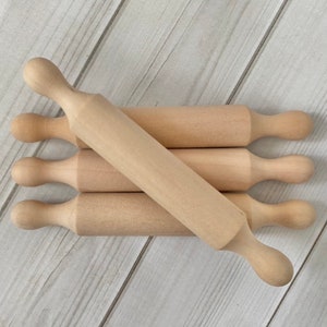 Puede incluir: Un conjunto de peque&ntilde;os rodillos de madera sin terminar. Cada rodillo tiene una superficie cil&iacute;ndrica y mangos redondeados. Los rodillos est&aacute;n dispuestos sobre una superficie de madera clara, sugiriendo un tema de reposter&iacute;a o cocina.