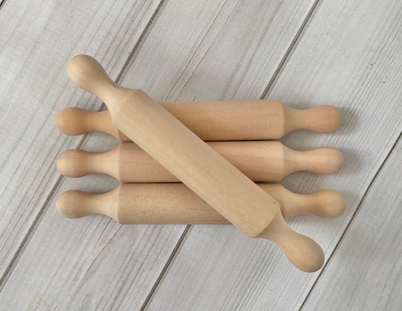 Puede incluir: Un conjunto de peque&ntilde;os rodillos de madera sin terminar. Cada rodillo tiene una superficie cil&iacute;ndrica y mangos redondeados. Los rodillos est&aacute;n dispuestos sobre una superficie de madera clara, sugiriendo un tema de reposter&iacute;a o cocina.