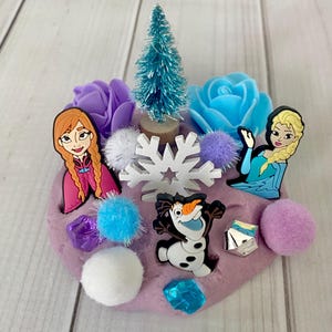 Könnte beinhalten: Eine lila Knetmasse-Kreation mit Figuren aus dem Film Frozen, darunter Anna, Elsa und Olaf. Die Szene ist mit einem kleinen blauen Weihnachtsbaum, Blumen, Pompons und Schneeflocken-Akzenten dekoriert.