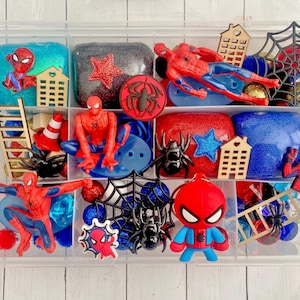 May include: A clear plastic container filled with various Spider-Man themed toys and craft supplies. Includes red and blue figurines, glittery blocks, wooden cutouts, spider webs, and decorative elements. The color scheme is primarily red, blue, and black.