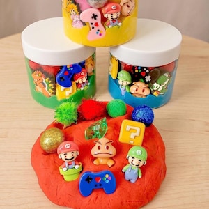 Puede incluir: Tres frascos de plástico transparente con tapas blancas llenos de slime de colores, pequeñas figuras de personajes de videojuegos y pompones. Al frente, una masa de slime rojo decorada con figuras de Mario y Luigi y un bloque.