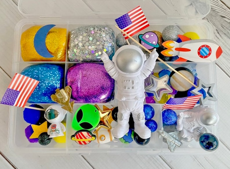 May include: A clear plastic storage container filled with various space-themed play dough and toys. The container has six compartments, each filled with a different color of play dough. There are also small astronaut figurines, stars, planets, and a rocket ship.