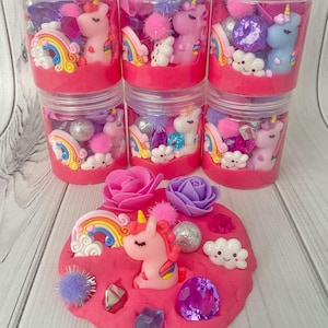 Peut inclure: Kits de jeu sensoriel sur le thème des licornes. Chaque kit comprend un pot en plastique transparent rempli d'éléments décoratifs roses, violets et argentés, et une petite figurine de licorne. Une pâte à modeler rose est décorée d'une licorne, de pierres précieuses et de fleurs.