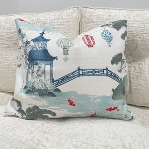 May include: A white pillow with a blue and green Asian-inspired design. The pillow features a pagoda, a bridge, lanterns, and fish.