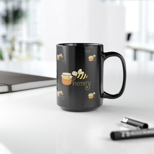 May include: A black ceramic mug featuring a repeating pattern of small cartoon bees and honey jars. The word honey is written in a cursive script font. The design includes yellow and orange accents against the solid black background of the mug.