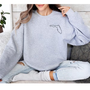 Puede incluir: Una sudadera gris claro con un contorno negro del estado de Florida en la parte superior izquierda del pecho. La sudadera de manga larga con cuello redondo está hecha de un material suave.