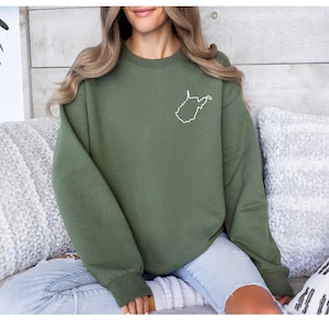 May include: An olive green sweatshirt featuring a white outline of the state of West Virginia on the upper left chest. The sweatshirt is a solid color and has a crew neck.