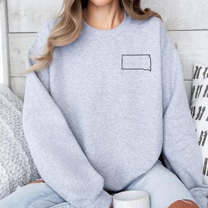 May include: A light gray sweatshirt featuring a black outline of the state of South Dakota. The sweatshirt is a crew neck style and has long sleeves. The state outline is located on the upper left chest area of the sweatshirt.